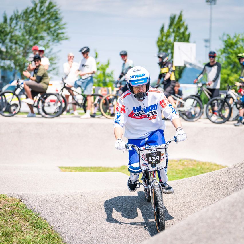 Ein Radfahrer fährt auf einer Rennstrecke ein kleines Fahrrad mit der Nummer 139, während andere dahinter zuschauen.