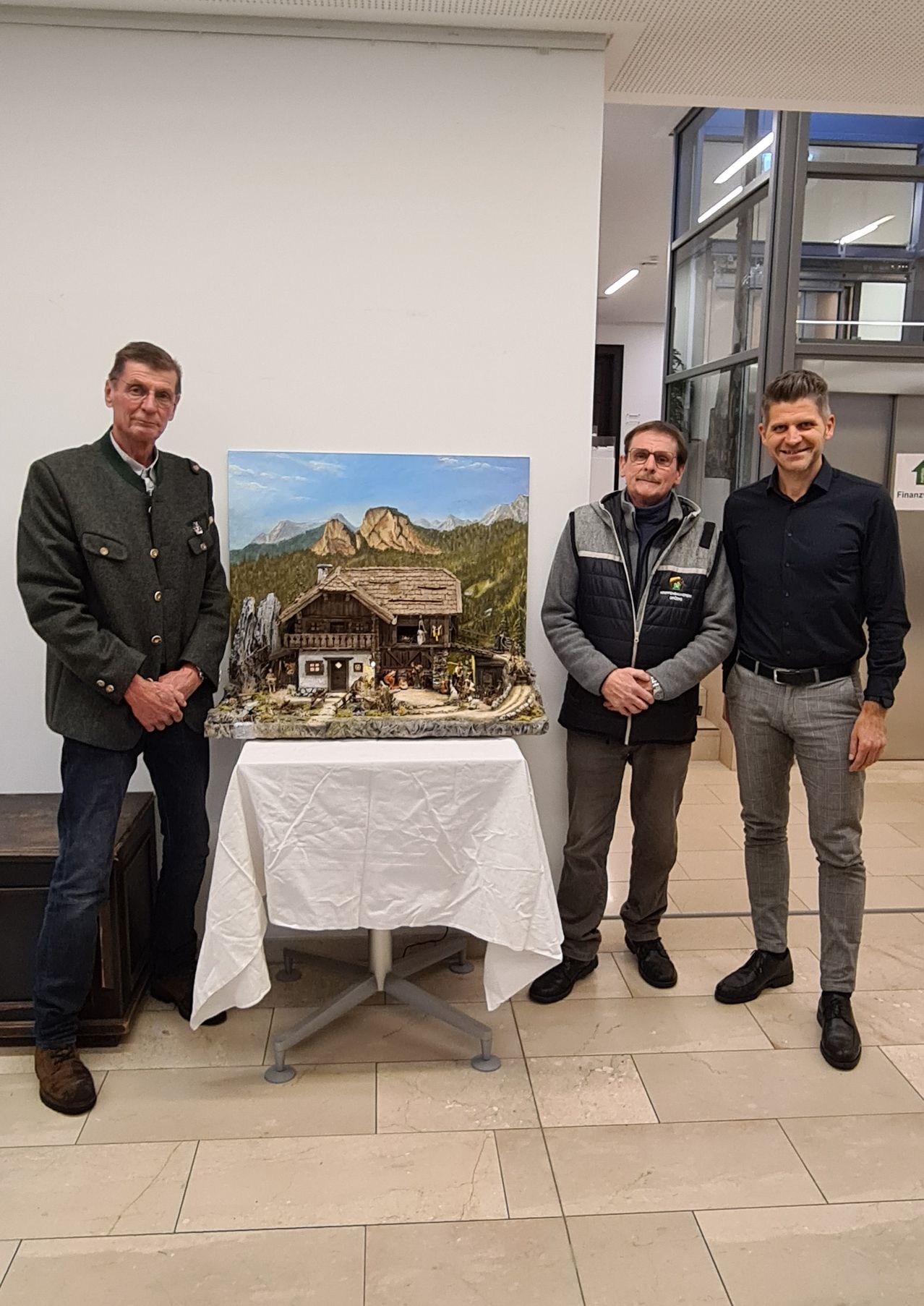 Drei Männer stehen neben einem Tisch mit einer detaillierten Landschaftsmalerei, die ein Haus und Berge zeigt, mit einer weißen Tischdecke.