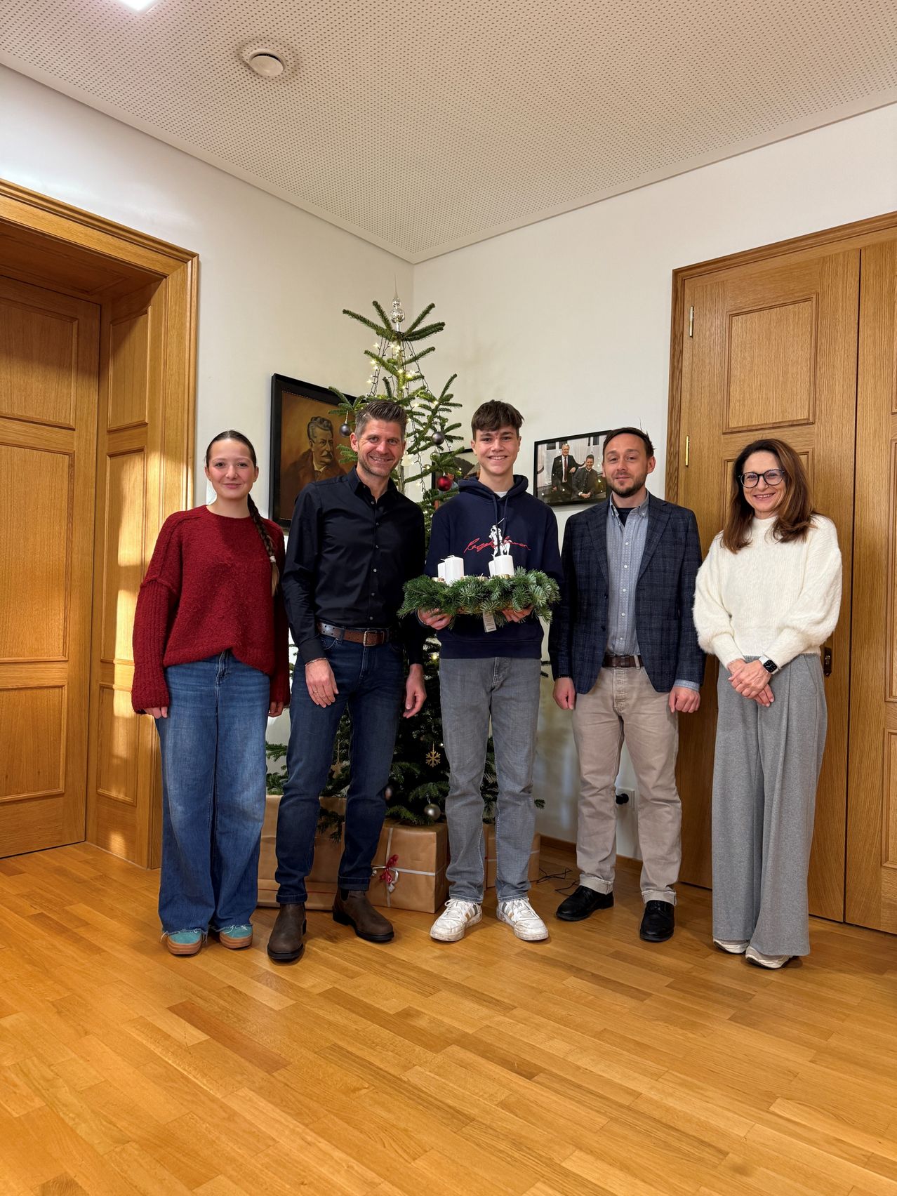 Fünf Personen posieren für ein Foto in einem Raum mit Holzböden und Türen. Ein Mann hält einen Weihnachtskranz mit brennenden Kerzen. Hinter ihnen steht ein Weihnachtsbaum und ein gerahmtes Bild an der Wand.