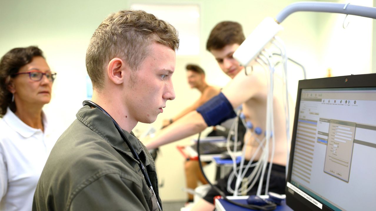 Ein Mann in einer Jacke und mit Stethoskop überwacht einen Patienten, der sich einer medizinischen Untersuchung unterzieht, in einem Raum mit medizinischen Geräten.