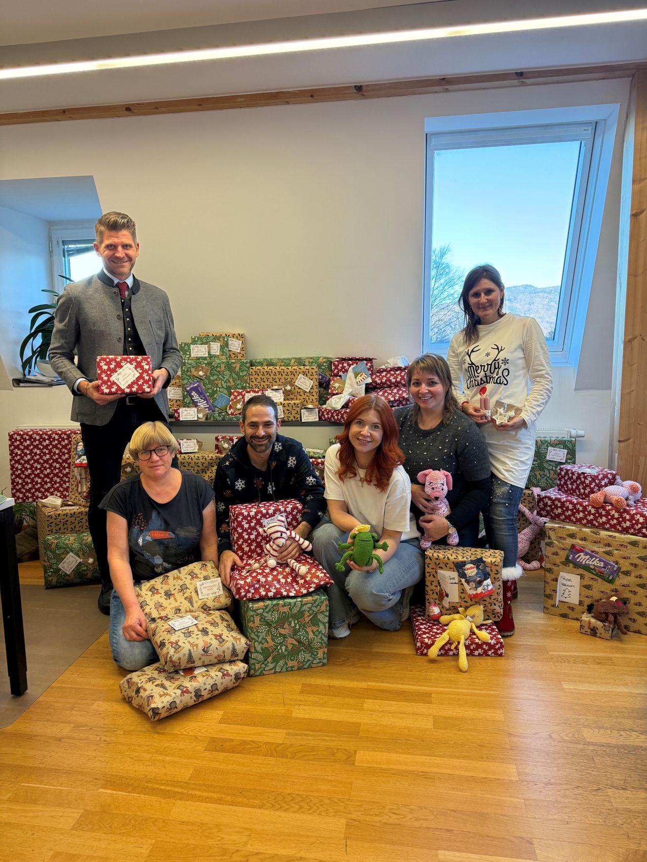 Mehrere Personen posieren mit Geschenken in einem Raum mit Holzböden. Ein Mann steht und hält ein Geschenk, während Frauen sitzen oder hockend Geschenke und Plüschtiere halten. Weitere Geschenke sind auf dem Boden und im Hintergrund angeordnet.