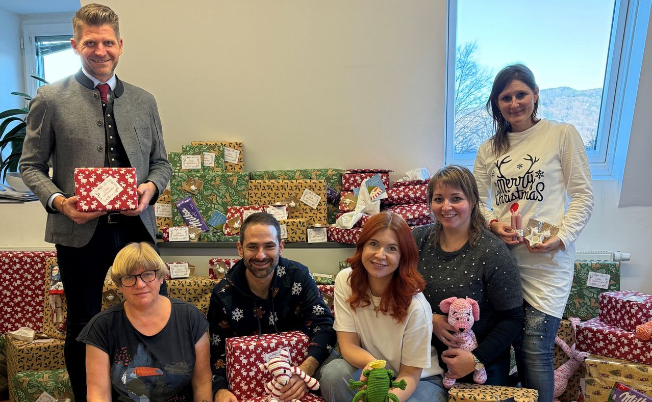 Eine Gruppe von Menschen lächelt und posiert für ein Foto mit Geschenken vor sich. Hinter ihnen stehen mehrere verpackte Geschenke auf dem Regal.