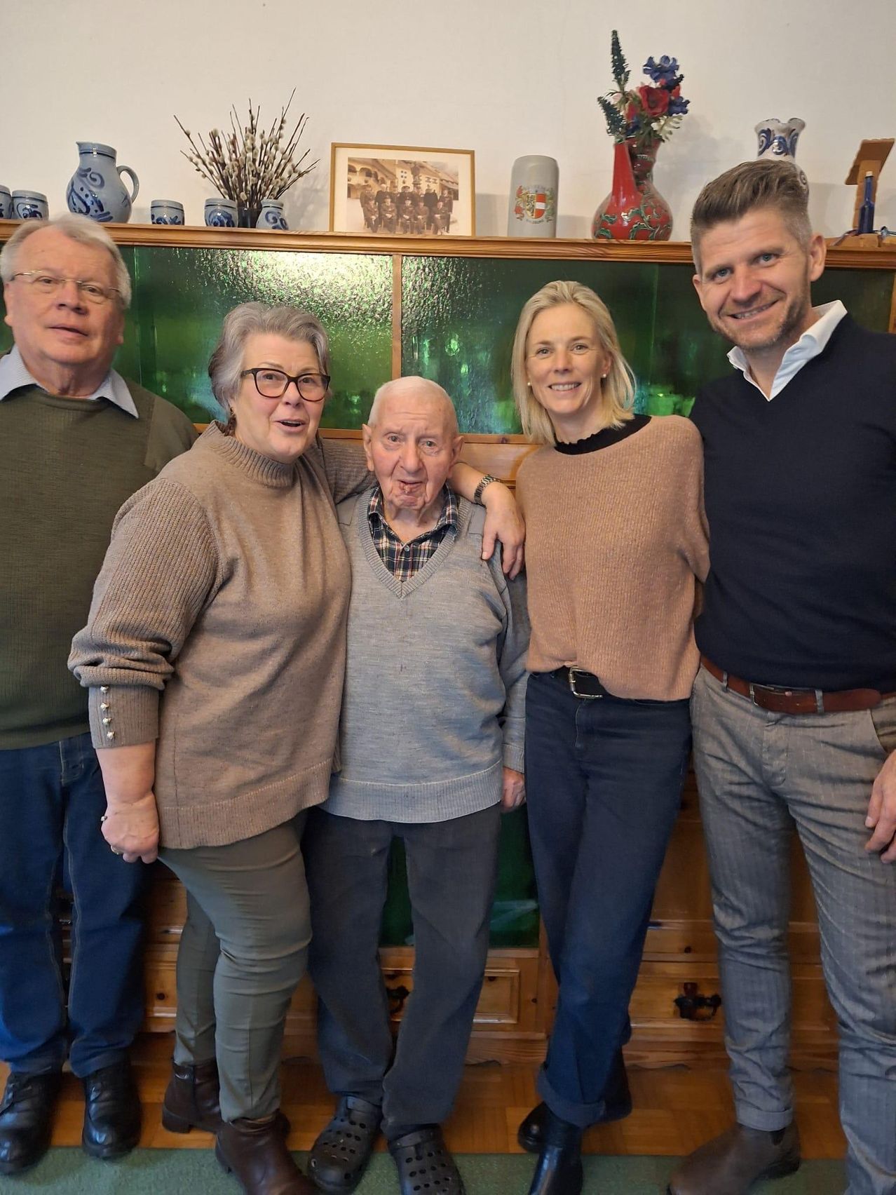 Fünf Menschen stehen zusammen in einem Raum und lächeln für ein Foto. Sie stehen vor einem Holzregal mit Dekorationsartikeln. Der Mann rechts trägt einen dunklen Pullover und einen braunen Gürtel.