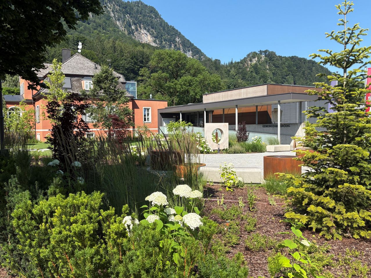 A modern building with a garden in front, surrounded by trees and plants. A mountain is visible in the background.