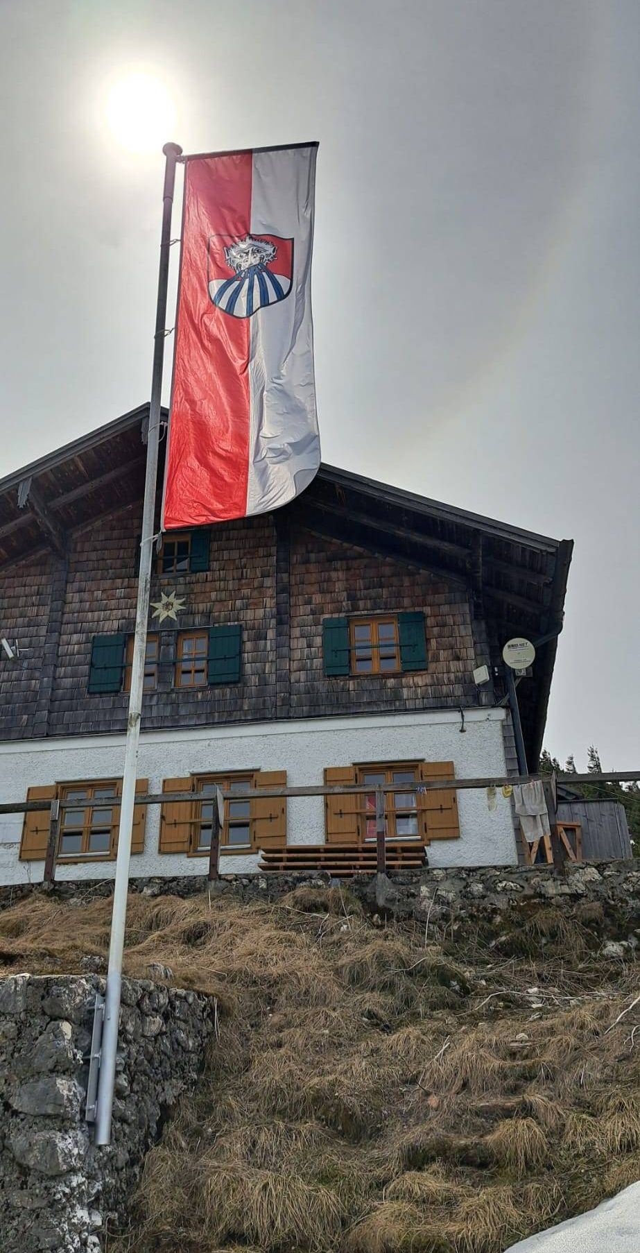 Ein traditionelles Haus mit einem Fahnenmast davor, der eine rot-weiße Flagge zeigt. Das Gebäude hat Holzläden und Fenster und eine Satellitenschüssel an der Seite.