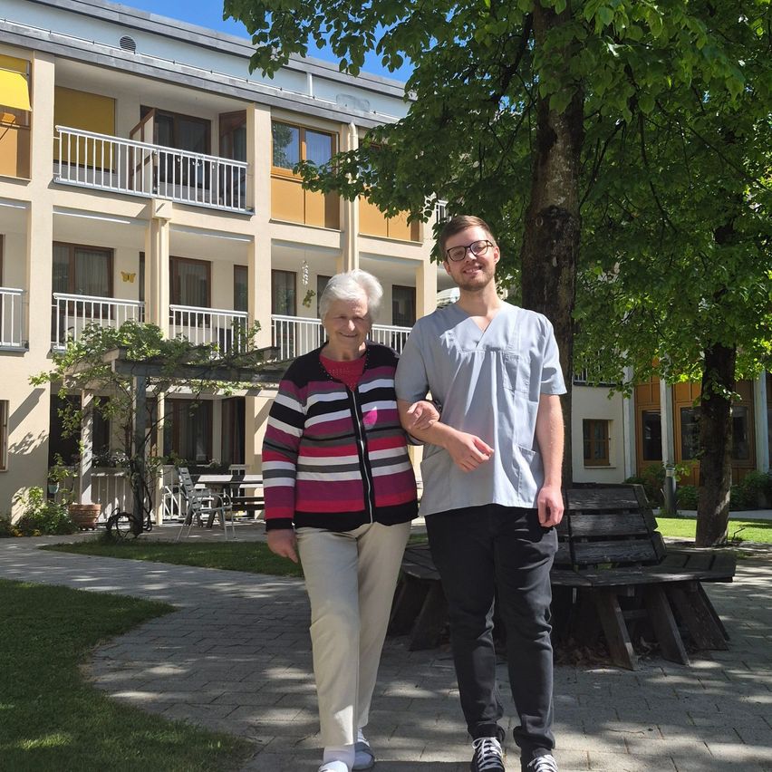 Eine ältere Frau und ein junger Mann stehen vor einem Gebäude mit Balkonen und einem Baum. Die Frau lächelt und der Mann trägt eine Brille.