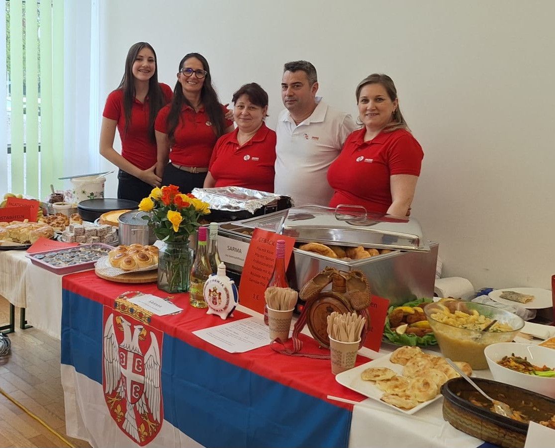 Fünf Menschen stehen hinter einem Buffettisch, geschmückt mit verschiedenen Speisen, Blumen und der Flagge Serbiens. Die Menschen lächeln, und ein Mann trägt ein weißes Hemd.