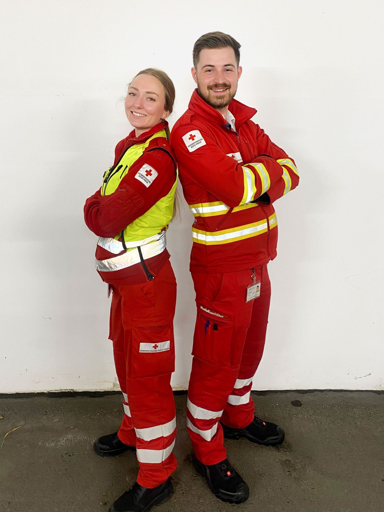 A smiling woman and a man stand together in red and yellow uniforms with red cross symbols. They are posing for a picture against a white wall.