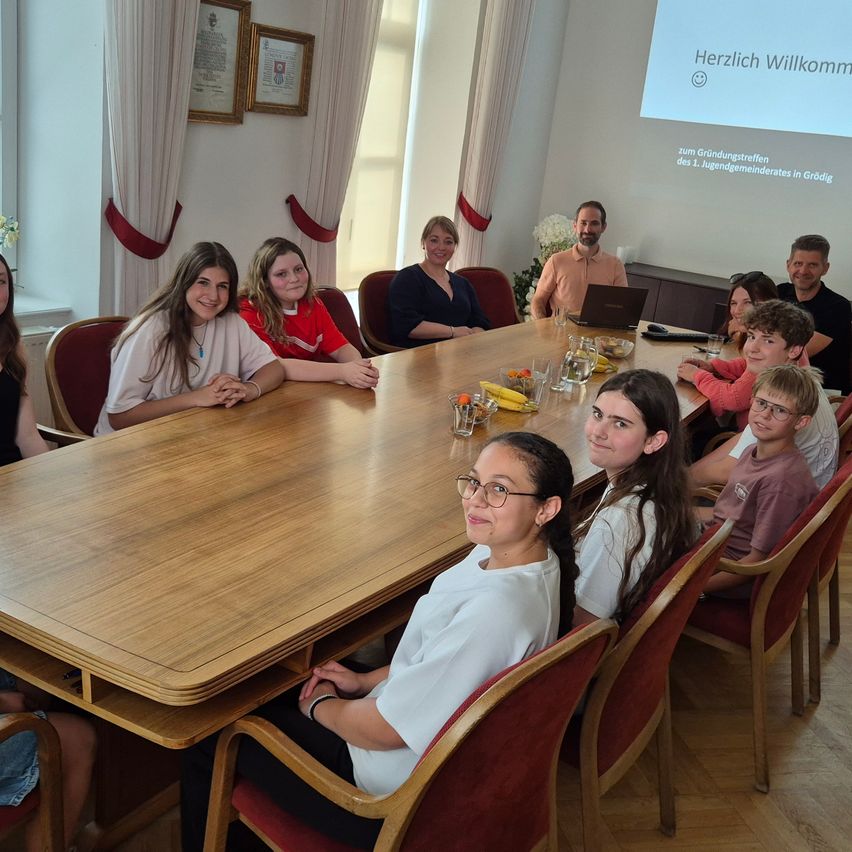 A group of people sit around a long wooden table in a room, smiling and posing for a photo. A projector screen displays a welcome message.