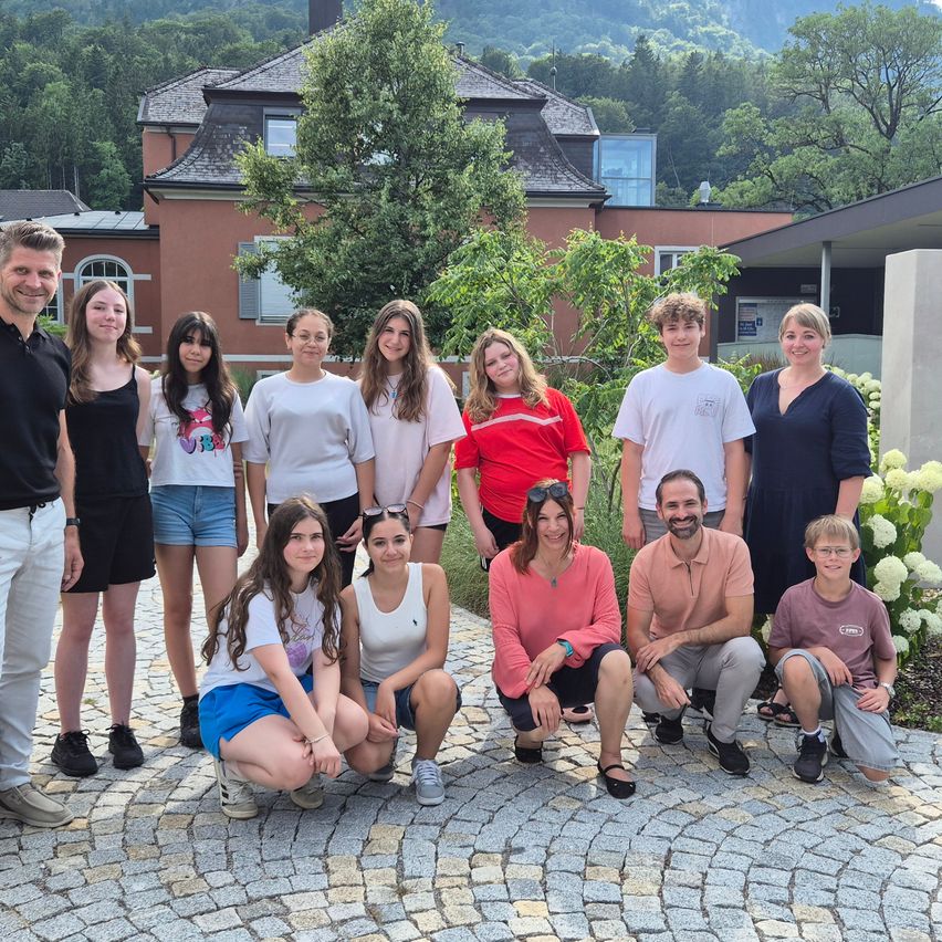 A group of people, including adults and children, are posing for a photo in front of a house with a garden. The house is surrounded by trees and mountains.