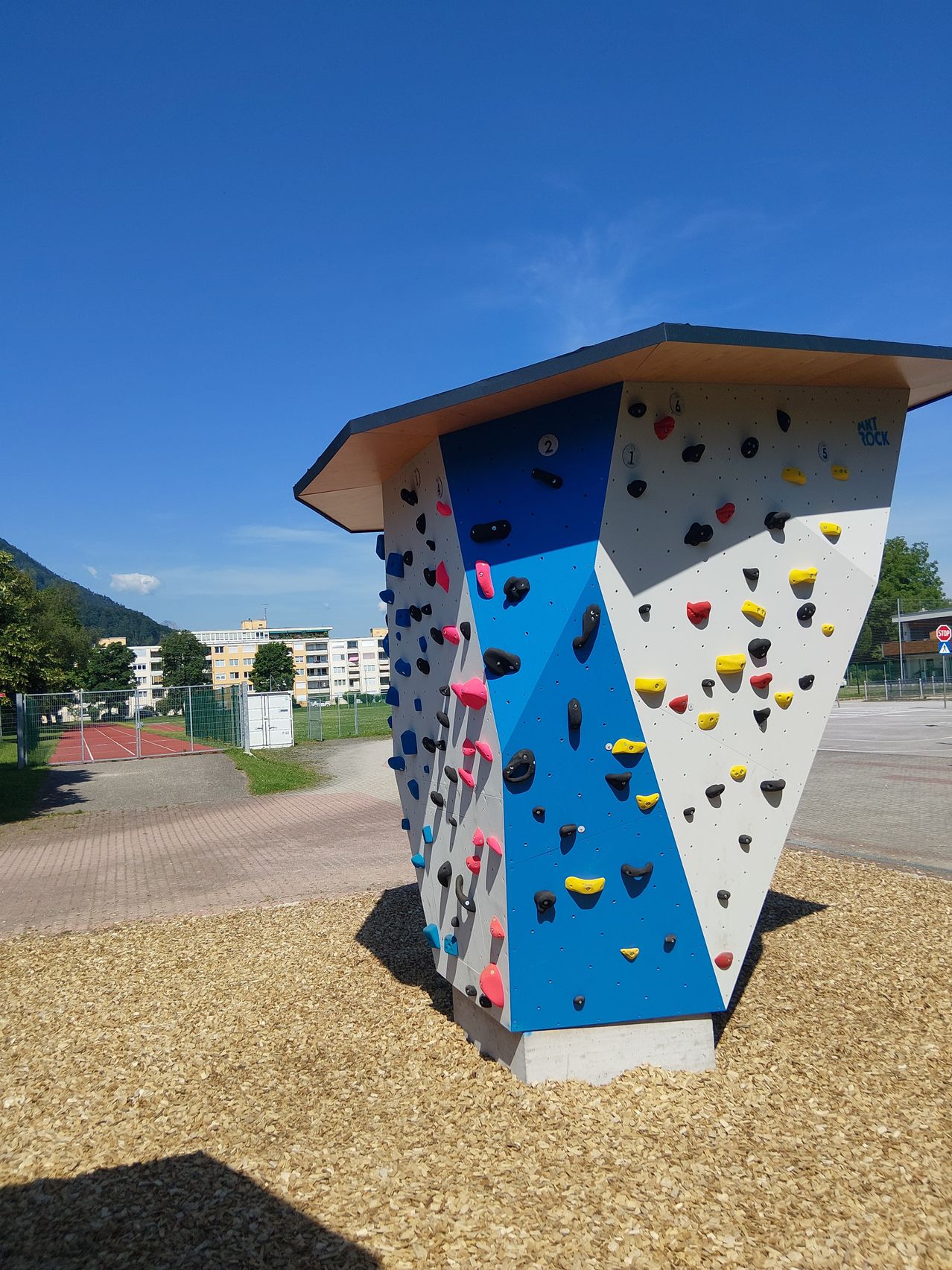 Eine bunte Kletterwand mit verschiedenen farbigen Griffen und einer dreieckigen Form steht in einem Park mit einem klaren blauen Himmel im Hintergrund.