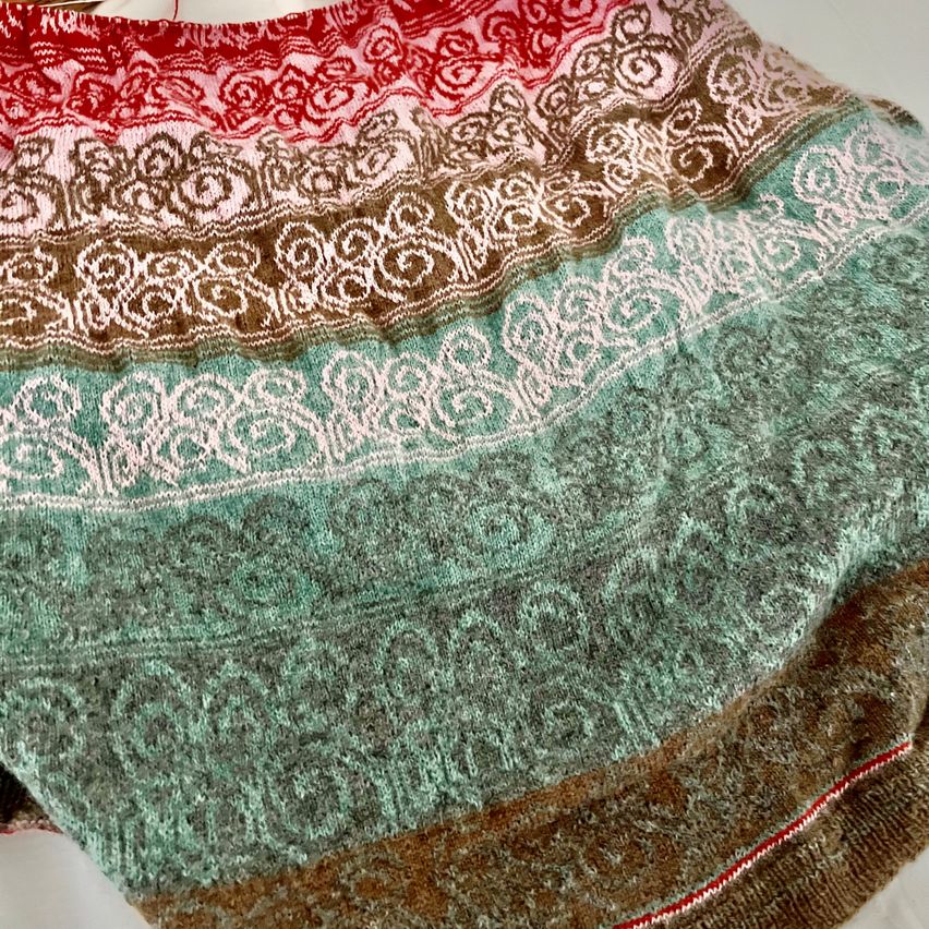 Ein Nahaufnahme eines gestrickten Stoffes mit einem mehrfarbigen Muster, das Rot-, Rosa-, Braun- und Grüntöne aufweist. Der Stoff hat ein kompliziertes Design mit Wirbeln und Blumenmotiven.