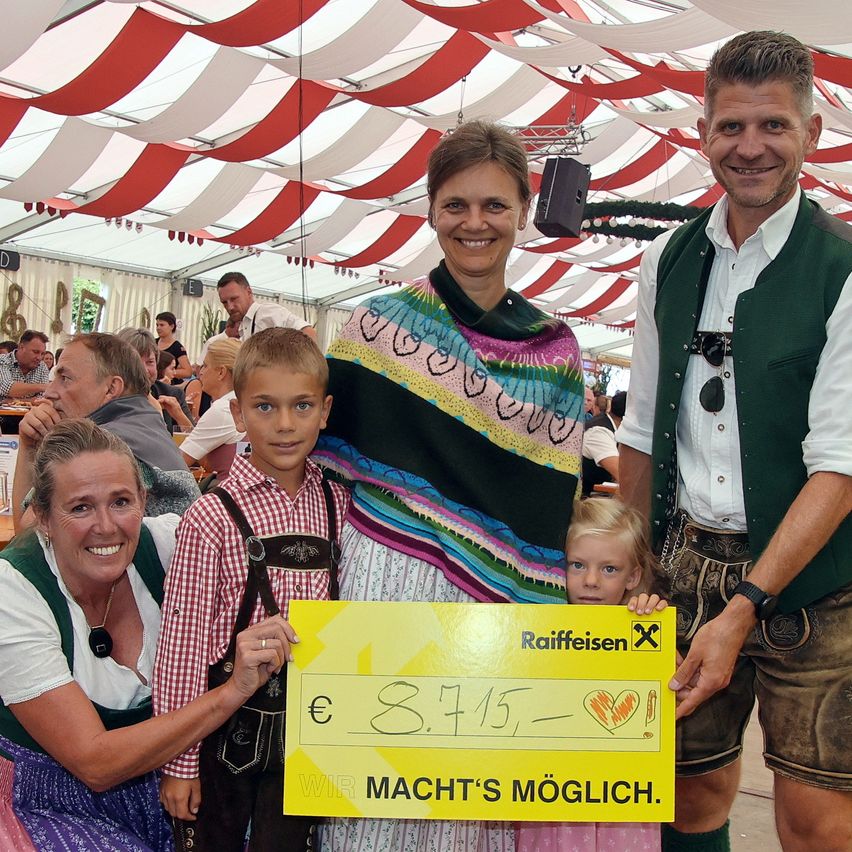 Eine vierköpfige Familie steht unter einem Zelt und lächelt für ein Foto. Der Mann hält einen gelben Scheck über 8.715 Euro. Die Frau trägt einen bunten Schal, und die Kinder tragen traditionelle Kleidung.