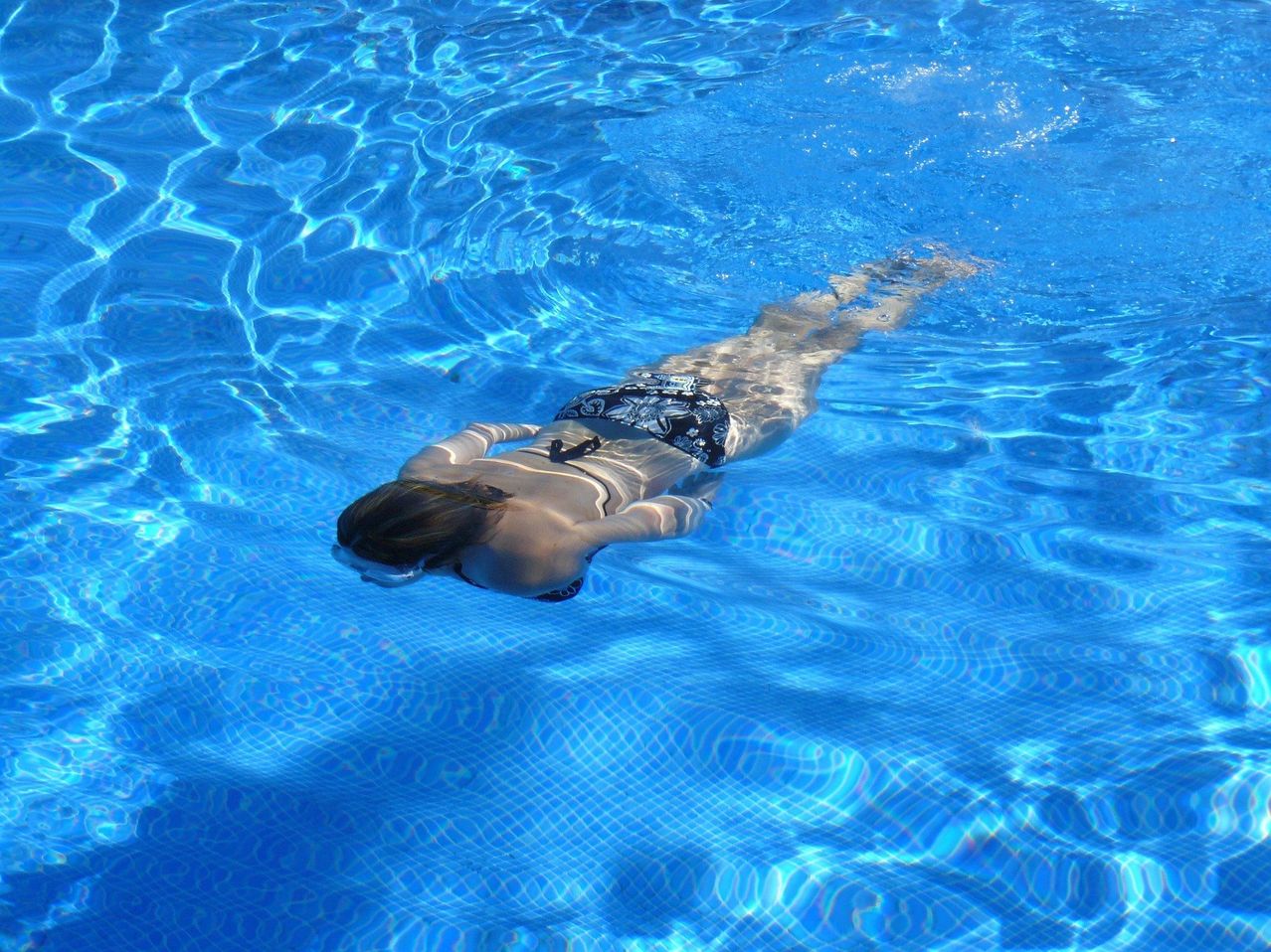 Eine Frau schwimmt unter Wasser in einem Pool, trägt einen schwarzen und weißen Badeanzug und eine Schwimmbrille. Das Wasser ist klar und blau.