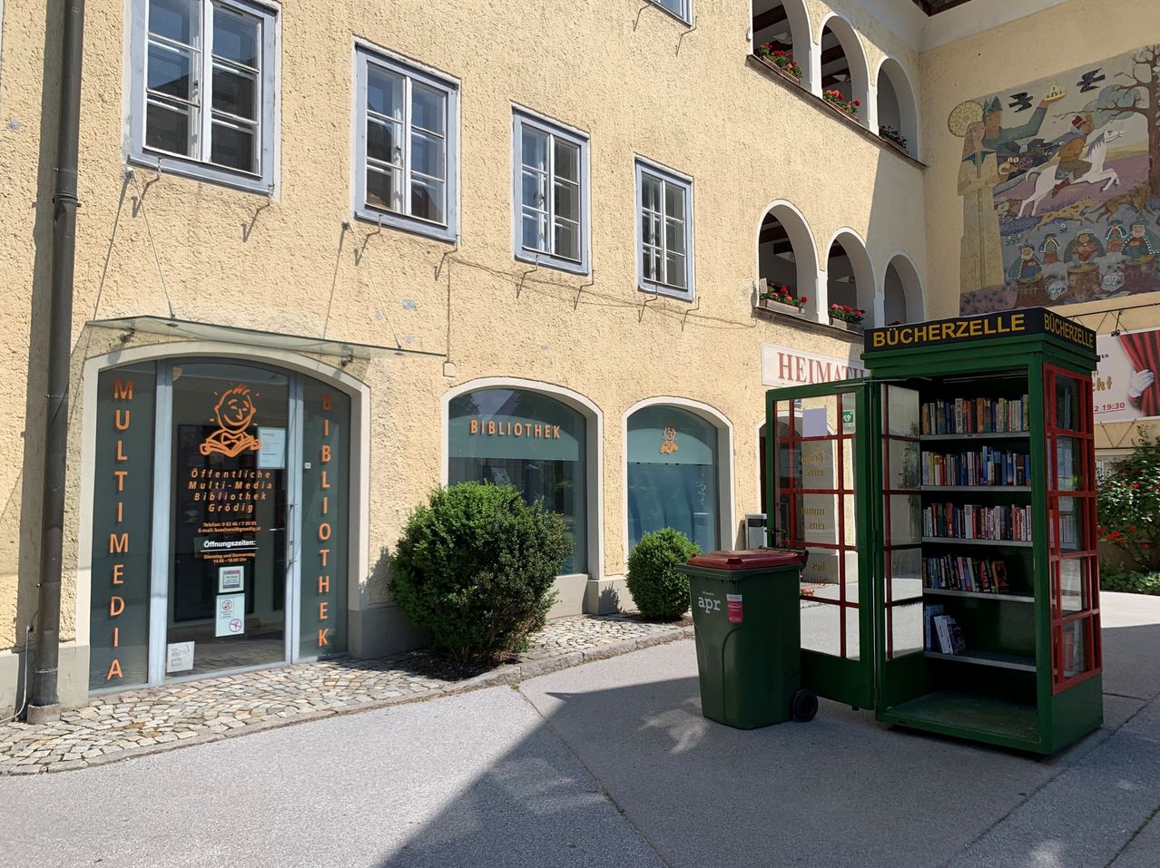 A beige building with several windows and arched doorways. A green trash bin is on the right, and a small green library box is on the sidewalk.