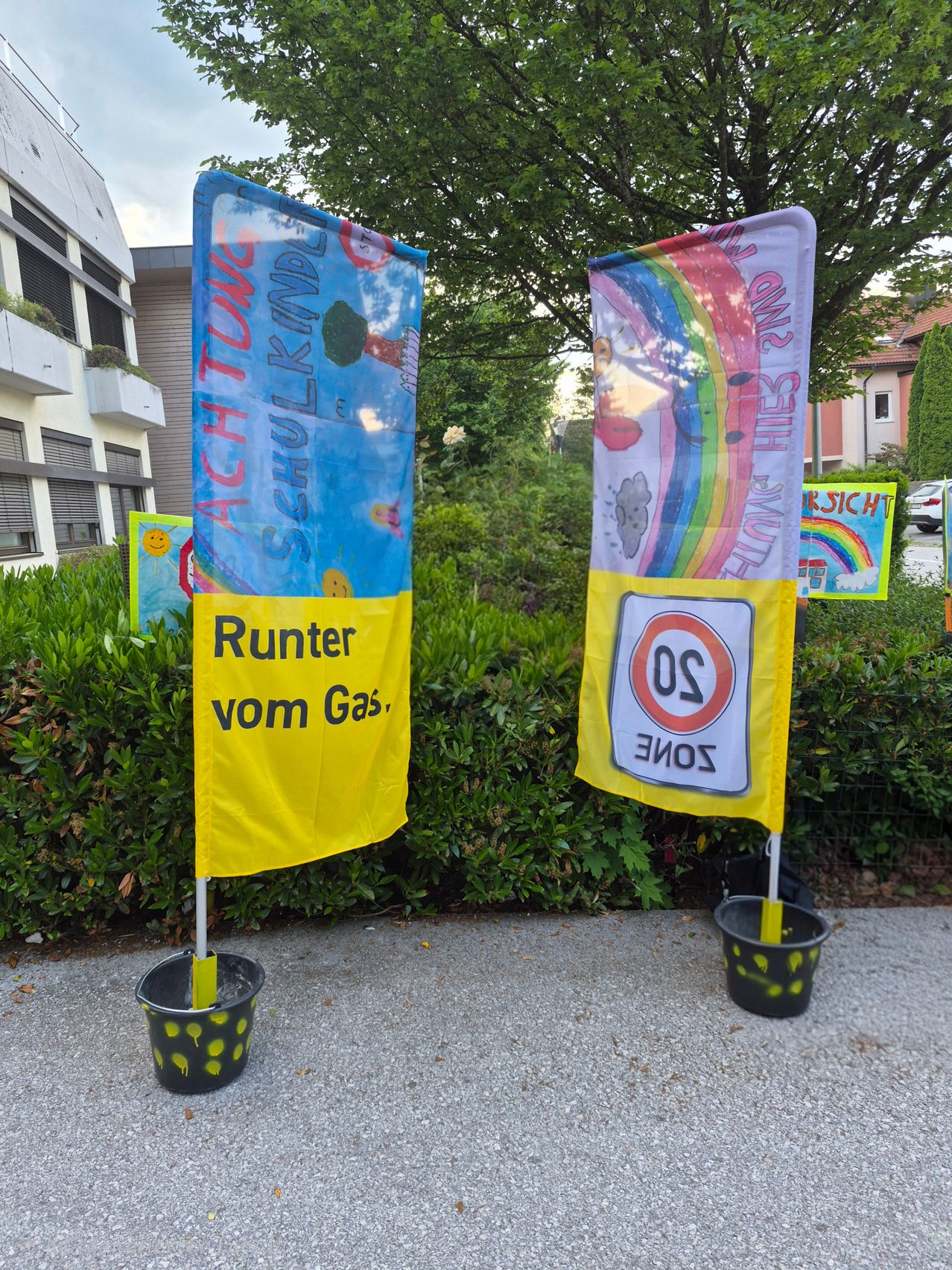 Two banners stand in front of a building. One has a rainbow and the word SOZIE, while the other reads 'Runter vom Gas'.