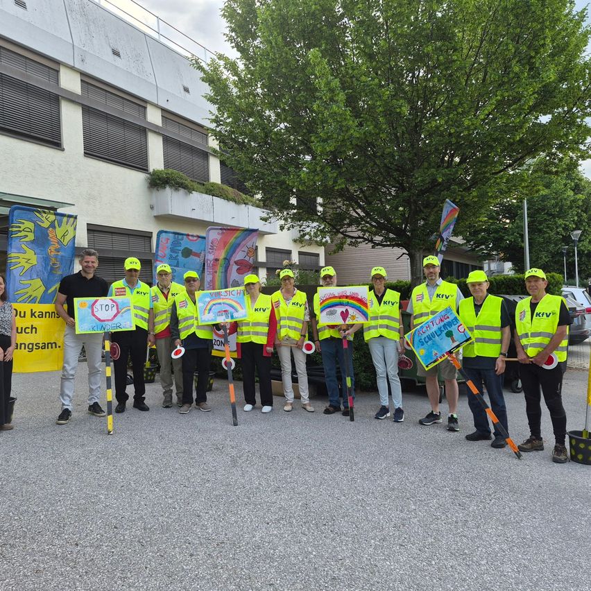 A group of individuals in yellow safety vests are standing outside, holding signs and flags, with a building and trees in the background.