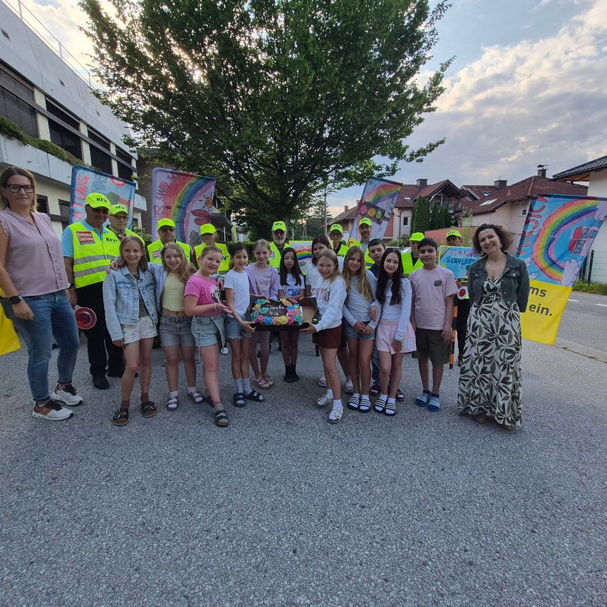 A group of children and adults stands outside, wearing yellow hats and vests, holding banners. They pose for a photo in front of buildings and a tree.