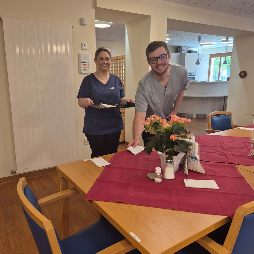 A man and woman stand at a table in a room, smiling and posing. The table has a red cloth, flowers, and condiments. Behind them is a kitchen with an open window.