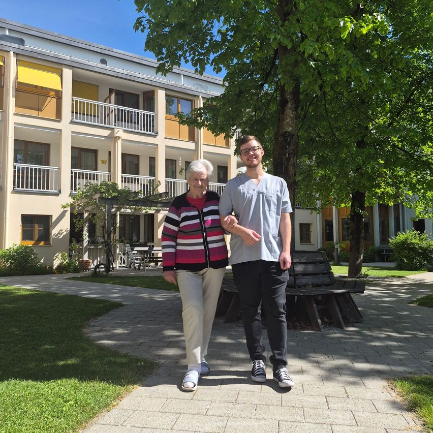 An older woman and a young man stand in front of a multistory building. They are smiling and posing for a photo.
