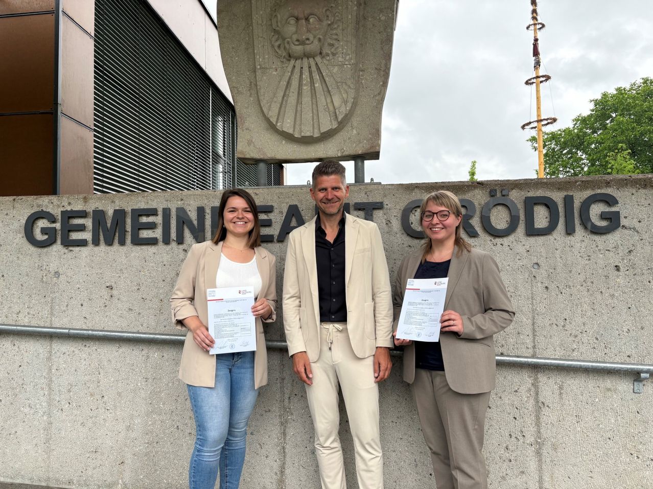 Three individuals stand before a concrete wall, smiling. They hold certificates in their hands. Behind them is a sign reading "Meine Heimat Crödel." There is a stone sculpture above the sign.