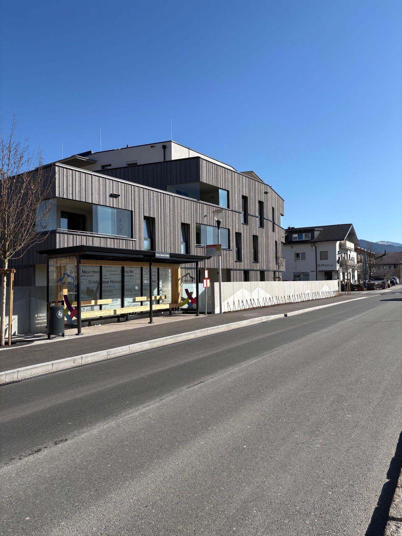 A modern building with large windows is on the side of a road with parked cars and trees under a clear sky.