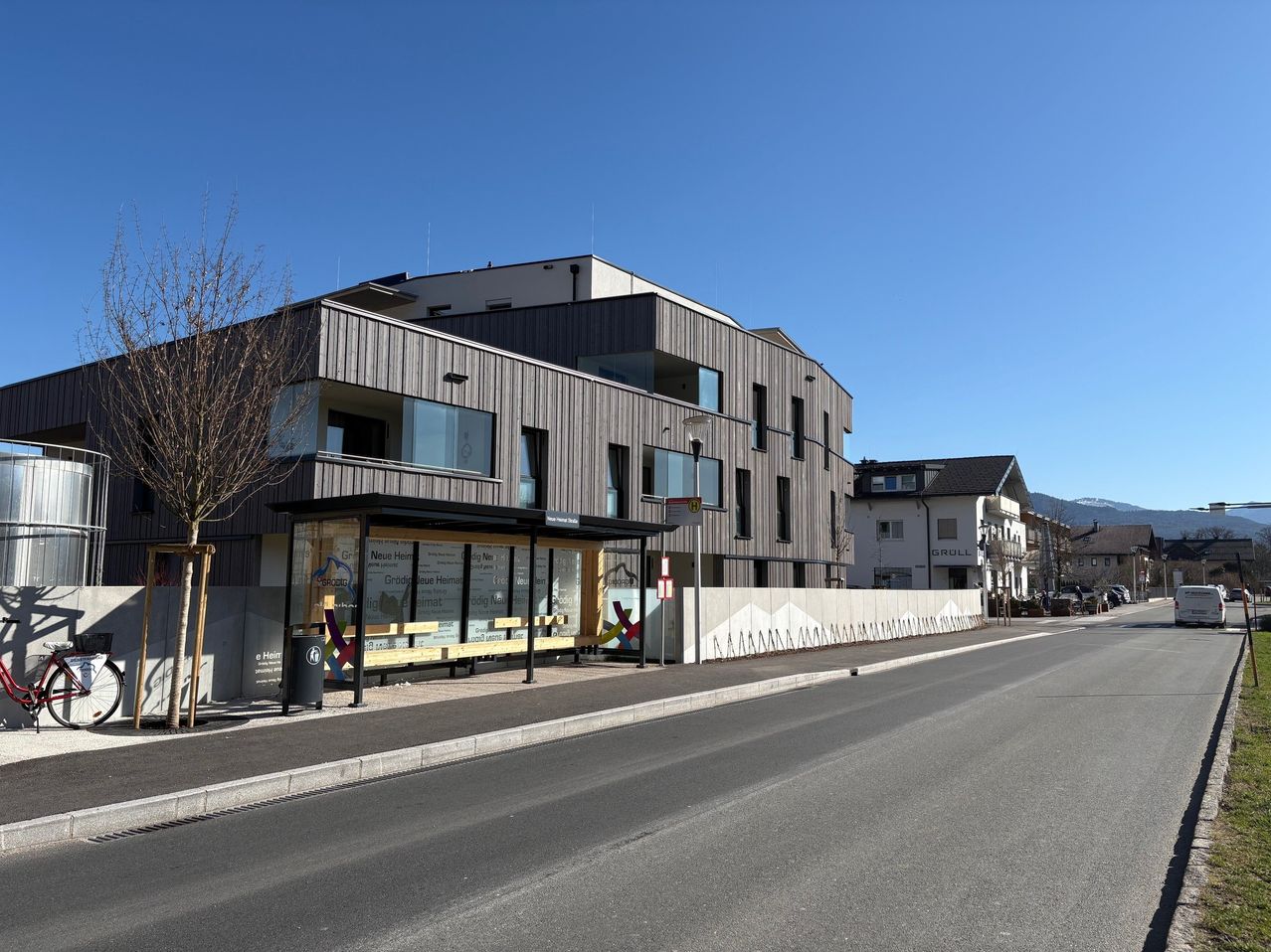 Eine leere Straße mit einem modernen Gebäude mit Glasfenstern. Eine Bushaltestelle mit farbenfrohen Werbungen ist davor. Ein Baum und ein Mülleimer stehen auf dem Bürgersteig.