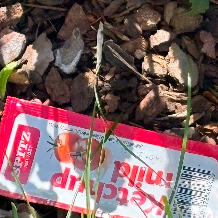 Ein rotes Paket mit der Aufschrift 'Spitz' für 'Margaritas' liegt auf dem Boden zwischen Steinen und Gras. Das Paket ist leicht zerrissen.