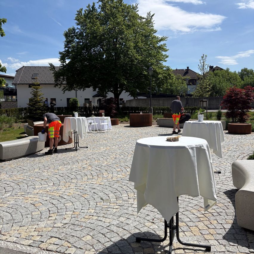Zwei Personen richten Tische in einem Park ein. Bäume und Büsche umgeben die Tische und ein Gebäude ist im Hintergrund.