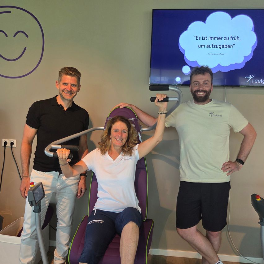 Three people are in a gym. One woman is sitting in a purple chair, holding a handlebar, and smiling. The two men stand beside her, smiling. Behind them is a large monitor displaying a smiley face and a motivational quote.