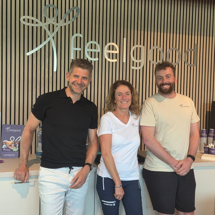 Three people stand in front of a counter. Two men and a woman are smiling. Behind them is a wall with the text Feelgood.