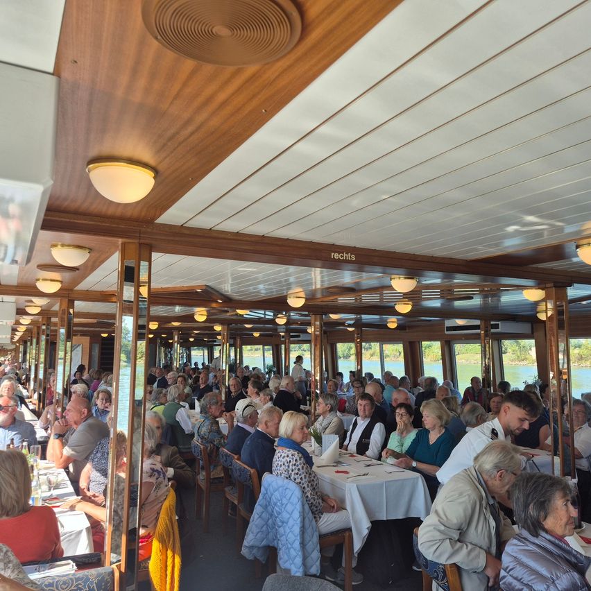Eine große Gruppe von Menschen sitzt an Tischen in einem Restaurant auf einem Boot. Das Restaurant hat Holzwände und Decken, mit mehreren Lampen, die von der Decke hängen. Die Tische sind mit weißen Tüchern bedeckt, und die Leute unterhalten sich.