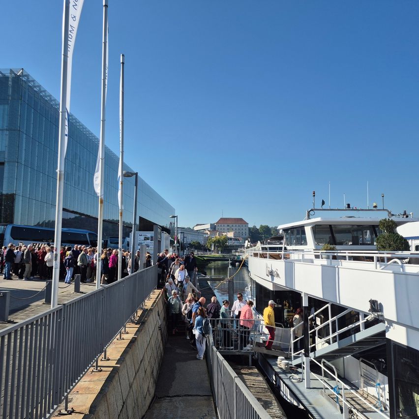 Eine Menschenmenge besteigt ein großes weißes Boot von einem Steg aus. Das Boot liegt in einem Hafen, mit einem modernen Glasgebäude im Hintergrund.