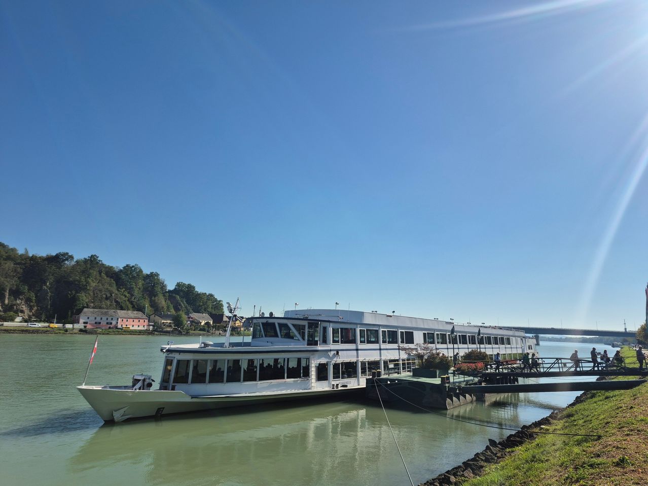 Ein großes weißes Kreuzfahrtschiff ist an einem Steg auf einem Fluss unter einem klaren blauen Himmel angedockt.
