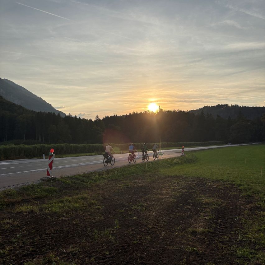 Fünf Radfahrer fahren auf einer Straße mit Bergen und Bäumen im Hintergrund, während die Sonne untergeht.