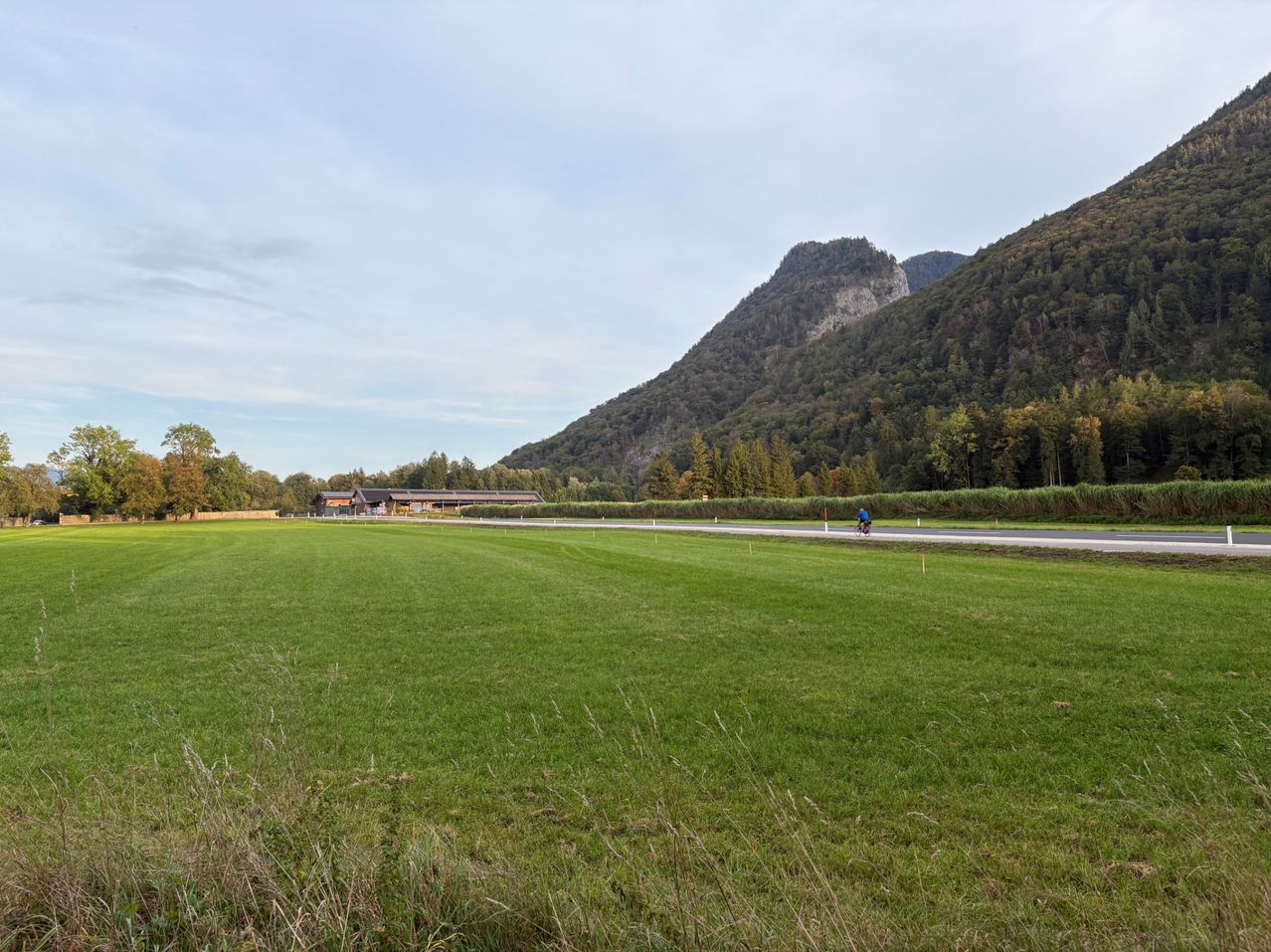 Ein weitläufiges, offenes Feld mit einer Straße, die hindurchführt. In der Ferne gibt es Berge, die von Bäumen bedeckt sind.