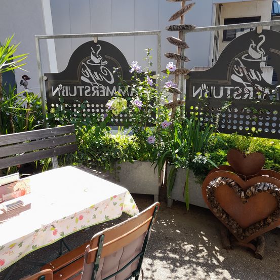 Eine Holzbank und ein Tisch mit einer blumigen Tischdecke stehen in einem Garten. Ein herzförmiger Pflanzgefäß ist in der Nähe. Hinter der Bank befindet sich ein Metallzaun mit einem Schild, auf dem 'Cafe Espresso' steht.