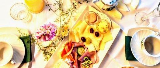 Ein Frühstückstisch mit verschiedenen Speisen, Getränken und Blumen. Es gibt zwei Teekannen, Weingläser, Teller und Tassen.