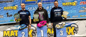 Drei Männer stehen auf einer Bühne und halten Modellautos. Sie haben Auszeichnungen und Trophäen. Ein Mann hält ein pinkes Auto, ein anderer ein blaues Auto und der dritte ein weißes Auto. Sie tragen schwarze Sweatshirts.