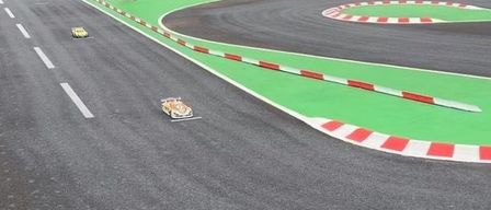 Eine Rennstrecke mit Menschen in der Nähe der Kurve. Zwei Autos sind auf der Strecke, und eines ist in der Nähe der Ziellinie. Es gibt Bäume und Büsche in der Umgebung.