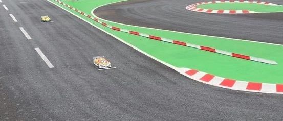 Eine Rennstrecke mit Menschen in der Nähe der Kurve. Zwei Autos sind auf der Strecke, und eines ist in der Nähe der Ziellinie. Es gibt Bäume und Büsche in der Umgebung.