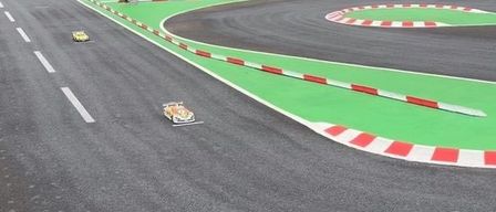 Eine Outdoor-Rennstrecke mit grüner und roter Strecke und mehreren Leuten, die herumstehen. Es gibt zwei kleine Autos auf der Strecke.