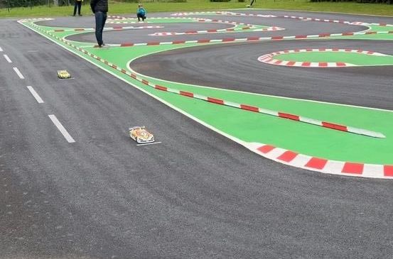 Eine Rennstrecke mit bemalten Markierungen, eine Person in Schwarz beobachtet zwei Spielzeugautos, während andere in Gelb und Blau stehen.