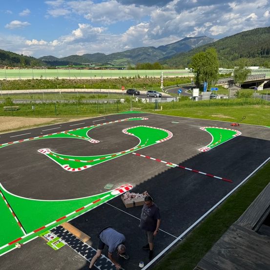 Zwei Männer malen eine Rennstrecke. Die Strecke ist grün und schwarz, mit weißen und roten Streifen. Ein Auto ist auf der Straße geparkt. Bäume und Berge sind in der Ferne.