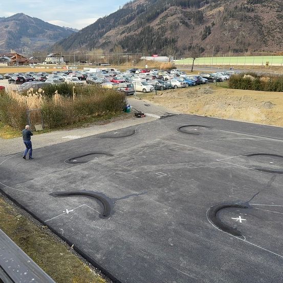 Auf einem Hügel geht ein Mann in der Nähe eines Parkplatzes mit vielen geparkten Autos, umgeben von Pflanzen und Bergen im Hintergrund.