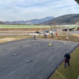 Eine Luftaufnahme einer im Bau befindlichen Straße mit Arbeitern, Bergen im Hintergrund und einer Brücke mit einer grünen Einzäunung.