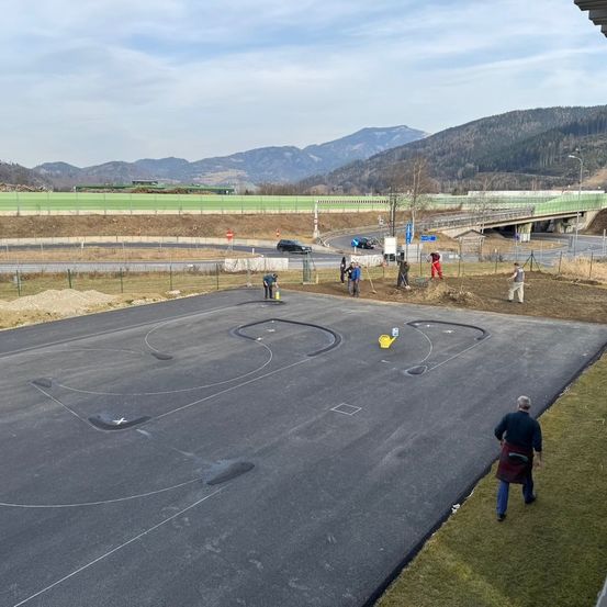 Eine Luftaufnahme einer im Bau befindlichen Straße mit Arbeitern, Bergen im Hintergrund und einer Brücke mit einer grünen Einzäunung.