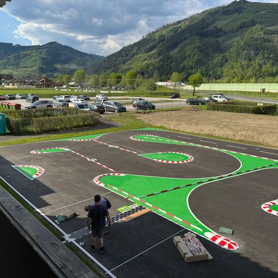 Eine Draufsicht auf eine Rennstrecke mit zwei Personen, die darauf laufen. Die Strecke ist von einem Zaun mit Büschen und Bäumen umgeben. Ein Parkplatz mit mehreren geparkten Autos ist in der Nähe. Berge sind in der Ferne.