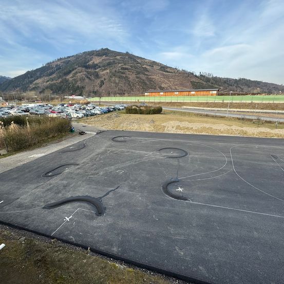 Ein großer asphaltierter Bereich mit einer Rennstreckenform, umgeben von einem Grasfeld und Bergen im Hintergrund. Ein großer Parkplatz ist in der Ferne zu sehen.