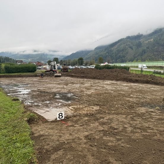 Ein Außenbereich mit der Nummer 8 auf dem Boden, umgeben von Erde und Gras, einem Baufahrzeug, Büschen und einem Berg in der Ferne.